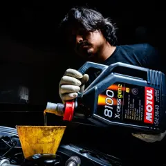 Mężczyzna w rękawiczkach przelewa olej silnikowy Motul 5W-40 z kanistra do silnika przez żółty lejek.