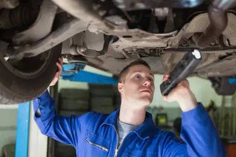 Mechanik w niebieskim kombinezonie bada podwozie auta, używając latarki. Sprawdza, jak wybić sworzeń wahacza.