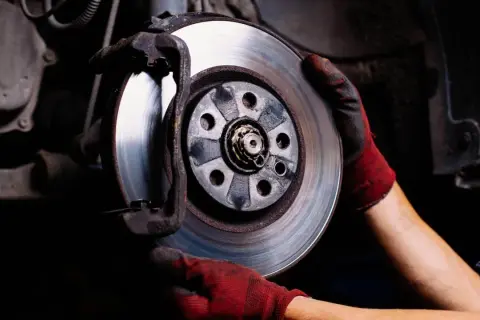 Mechanik w czerwonych rękawicach wymienia tarczę hamulcową. Hamulec ręczny trzyma jedno koło, zapewniając bezpieczeństwo podczas prac.