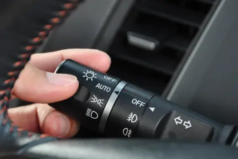 Dłoń manipuluje przełącznikiem świateł w samochodzie, ustawiając go na pozycję "AUTO".