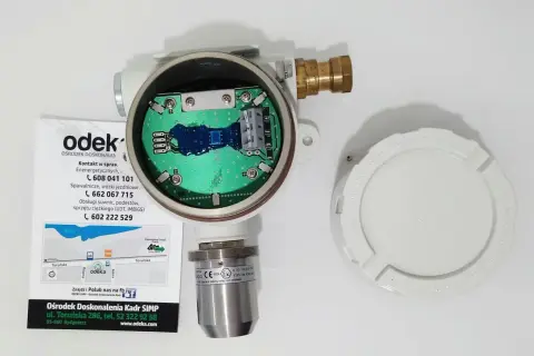 Otwarty detektor gazu z widoczną płytką drukowaną. Obok leży biała pokrywa z napisem "KEEP COVER TIGHT WHEN CIRCUITS ARE LIVE". To może być upg co to jest?