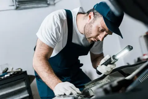 Mechanik w białej koszulce i granatowym kombinezonie, z latarką w ręku, sprawdza poziom płynu do sprzęgła w samochodzie.