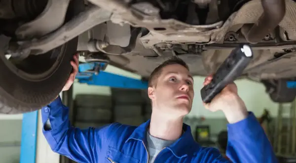 Mechanik w niebieskim kombinezonie bada podwozie auta, używając latarki. Sprawdza, jak wybić sworzeń wahacza.