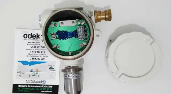 Otwarty detektor gazu z widoczną płytką drukowaną. Obok leży biała pokrywa z napisem "KEEP COVER TIGHT WHEN CIRCUITS ARE LIVE". To może być upg co to jest?