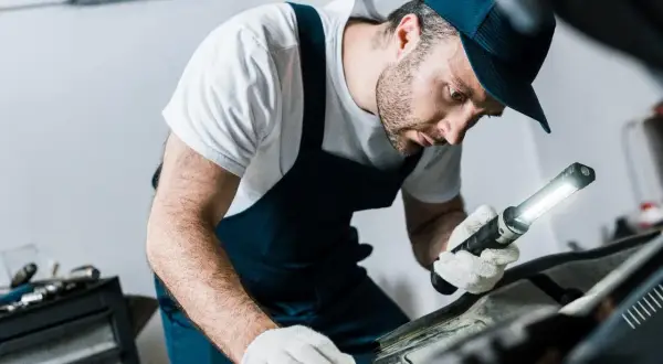 Mechanik w białej koszulce i granatowym kombinezonie, z latarką w ręku, sprawdza poziom płynu do sprzęgła w samochodzie.