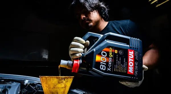 Mężczyzna w rękawiczkach przelewa olej silnikowy Motul 5W-40 z kanistra do silnika przez żółty lejek.