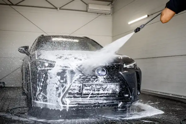 Czarny Lexus jest obficie spieniony podczas mycia na myjni. Widać strumień wody pod ciśnieniem, który spływa po masce i grillu.