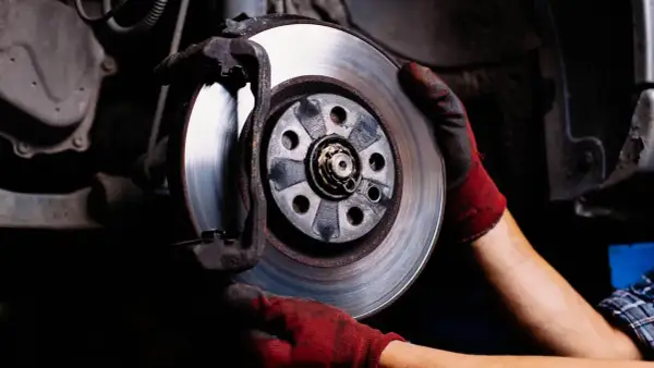 Mechanik w czerwonych rękawicach wymienia tarczę hamulcową. Hamulec ręczny trzyma jedno koło, zapewniając bezpieczeństwo podczas prac.