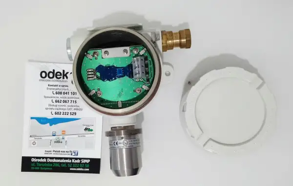 Otwarty detektor gazu z widoczną płytką drukowaną. Obok leży biała pokrywa z napisem "KEEP COVER TIGHT WHEN CIRCUITS ARE LIVE". To może być upg co to jest?